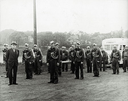 A black and white photograph of a group of men outside, standing in rows, wearing mine rescue gear. They each have a pack on their backs, with tubes going to a face mask resting below their necks and wearing a hat with a light on the front. A man in a suit is standing in front of the group, looking at the camera. The photo is circa 1943.