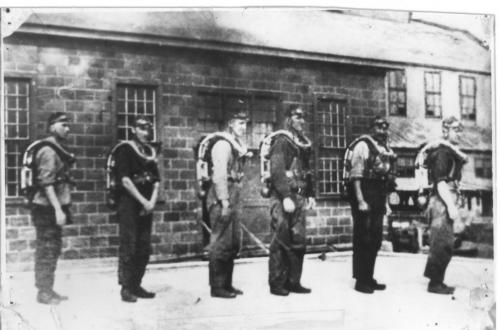 : A black and white photograph of a group of 6 men, turned sideways, to better show the breathing apparatus each was wearing so that they could work safely in potentially poisonous air in a coal mine after an explosion. They were the B team of the Acadia Rescue Corps, circa 1924.