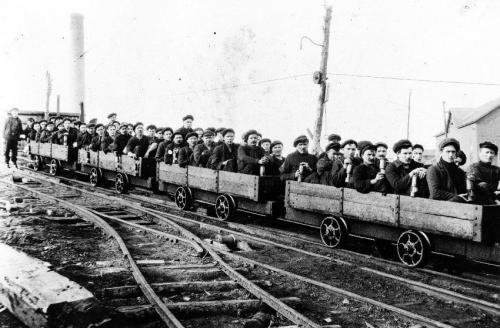 : short-sided carts on rails hold several miners and are attached in a row of 4 carts, about to descend into a slope mine. The cart is called a rake.