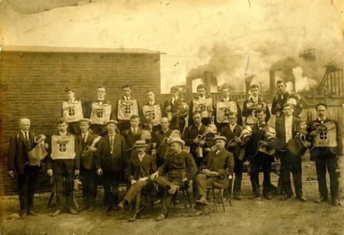 A sepia-toned photograph of a large group of men. Most are wearing a protective chest plate or breathing bag, some are holding helmets, and others have pieces of equipment. They are wearing suits. Three men are sitting in front, all wearing hats. In the background are several smokestacks billowing black smoke.