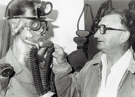 A black and white photograph of draegerman Robert Hoegg pointing out the pieces of his mine rescue equipment on a mannequin, circa 1980. The equipment included a mine helmet with light, goggles, an oxygen mask over the mouth, a nose clip and tubes leading to a pack that is not in the photo.