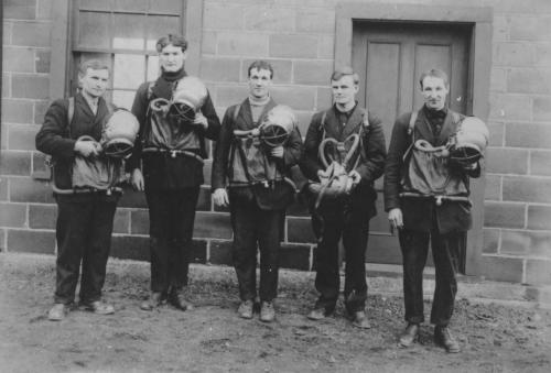 An undated black and white photograph of a group of 5 men wearing suits and their draeger equipment. They have breathing bag pouches in front with breathing tubes attached and are holding metal helmets.