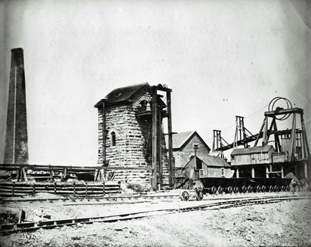 A black and white photograph of some of the buildings at the Foord Pit. It shows a smokestack to the left, and in the center is a sandstone building, called a Cornish pumphouse, which enclosed the pumping mechanism that pumped water out of the mine. To the right are other buildings and wheels and rope of the mine hoist. In front is a row of pit tubs.