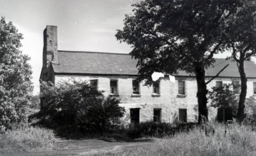 A photograph of a very British-looking, large, two-storey building. Eight windows are visible on the second storey. Shrubs block part of the building.