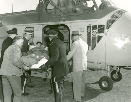 A black and white photograph of two RCMP officers and a few other men handling a stretcher containing a man wrapped in blankets being loaded into the open door of a helicopter.