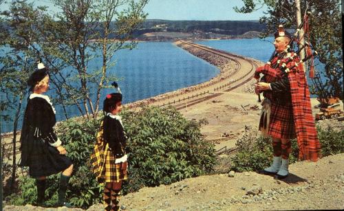 A colour postcard of the Canso Causeway in the background, with a Scottish bagpiper playing the pipes and two girls in Scottish kilts and tams on a hill above. This was a common Cape Breton tourism promotional image in the 1960s. The postcard title is “The Road To The Isles Causeway”.