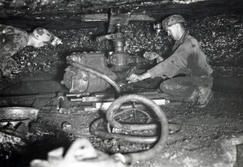 A black and white photography circa 1940 of a miner crouching beside a coal shearer, hands on the controls, roof close to his head.