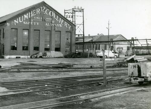 a black and white photograph of a building on the site of the Number 12 Colliery, New Waterford. Words painted on the end of the building read Number 12 Colliery Since 1908 Safety Always.