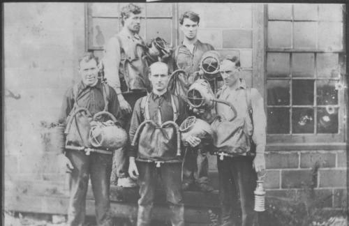 n undated black and white photograph of a group of five unidentified early Pictou County draegermen. They are wearing large pouches in front, held on with straps around their shoulders with two breathing tubes coming from the pouch. Each man is holding a metal helmet, similar to the type used for deep-sea diving. One also is holding a miner’s lamp.