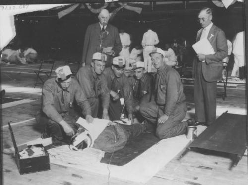  An undated black and white photograph of a mine rescue competition. A group of men all dressed alike are on their knees attending to a man laid out on the floor. They have bandaged his head, arm and leg. In the background can be seen other groups engaged in similar activities.