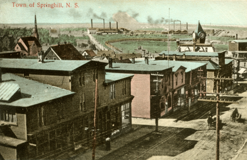 A hand-coloured postcard of the buildings on the main downtown street of Springhill, with the smokestacks and buildings of two coal mines seen in the distance. The postcard is undated.