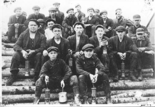 a black and white photograph of a group of 22 Vale Colliery miners from Thorburn. It includes both men and boys, sitting on a pile of logs that will be used for pit timbers.They are holding mine lamps and piece cans - which are lunch buckets - circa 1910.