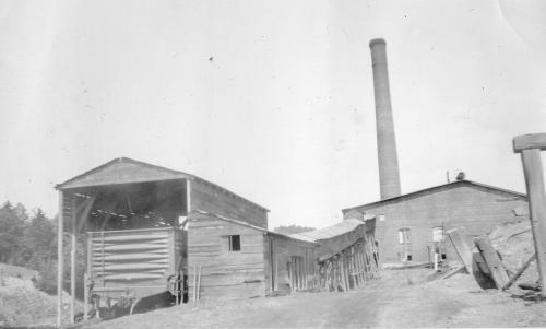 Black and white photograph showing buildings of the Victoria Mine in River Hebert, including a rail car in an enclosure, a covered long, narrow building on stilts (possibly to protect a conveyor or mine trip) and a smokestack behind a non-descript building. The photo is undated.