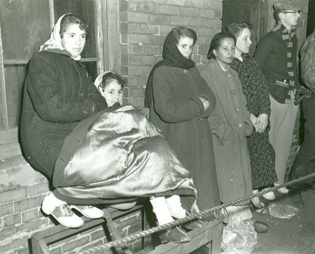 A black and white photograph of a group of 5 women and 1 man in coats leaning against a brick building. One woman has another coat across her legs. They look chilled. They are waiting for word about family members trapped after an underground earthquake in Number 2 Mine, Springhill.