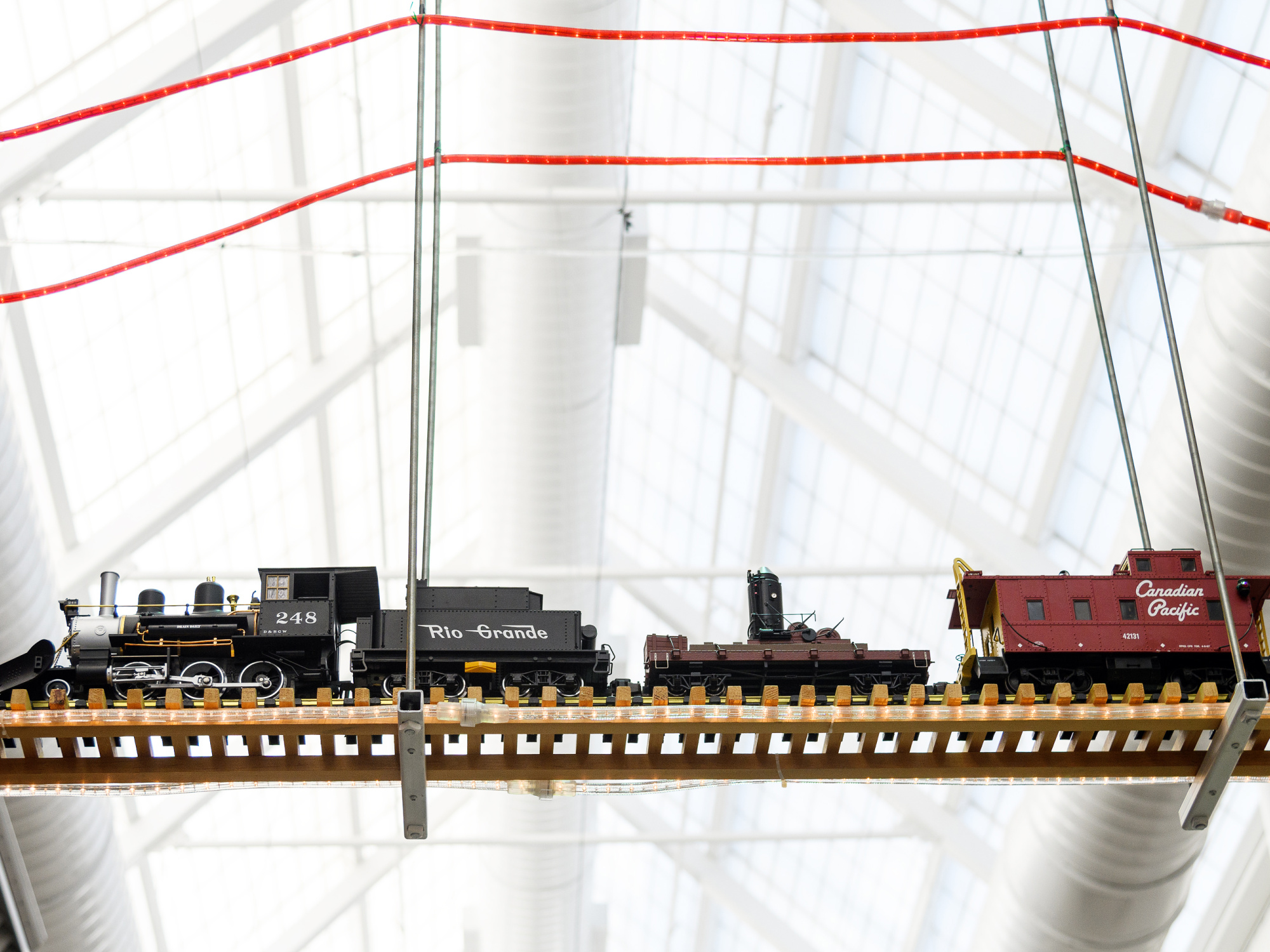A model train with a black locomotive labeled '248', a brown tender labeled 'Rio Grande', and a red caboose labeled 'Canadian Pacific', running on a wooden track. The train is suspended from a white ceiling with red and silver pipes in the background.