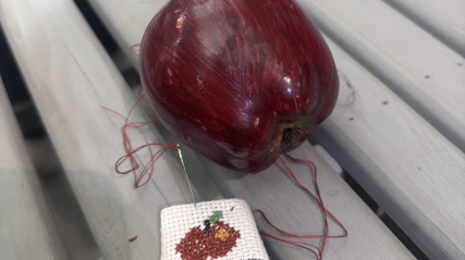 An image showing a high-angle view of a deep red artificial apple and a small cross-stitch project resting on a light grey, slatted wooden surface.
